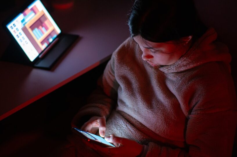 Woman looking tired and overwhelmed sits in a dark room, her face illuminated only by the glow of her laptop screen, symbo...